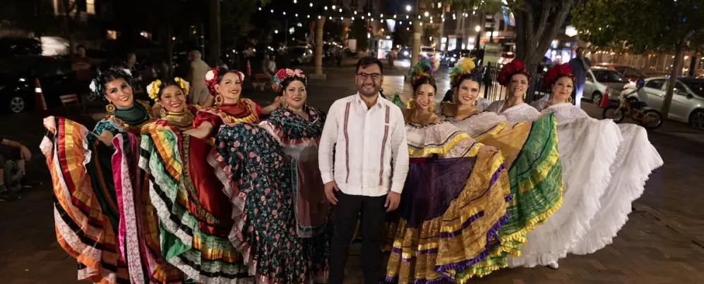 A group of folklórico dancers in colorful traditional dresses pose together outdoors at night during a cultural performance in Washington, DC.